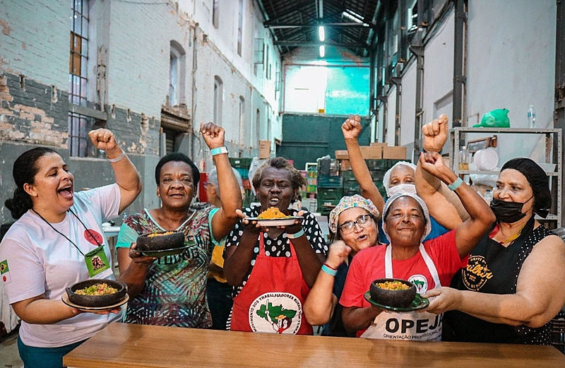 Foto de mulheres diversas segurando comida e com o punho levantado. Usam o avental do MST.