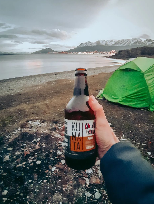 Foto da mão do Edson segurando uma garrafa de 500ml de cerveja chamada KUMATA. Ao fundo, parte de sua barraca verde acampada sobre um mar e um céu de inverno