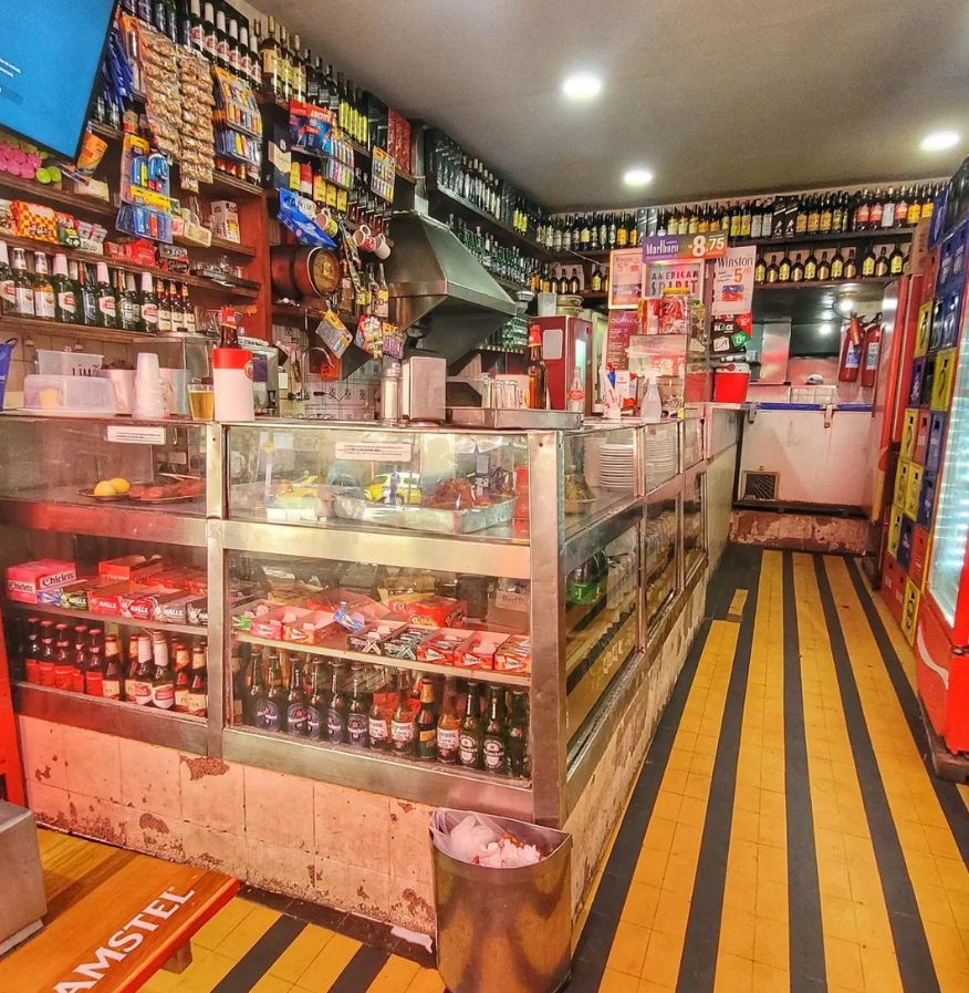 Foto de um butequim visto de fora. O local possui um chão com listras amarelas e verdes. Em suas paredes, diversos salgadinhos, pimentas, penduricalhos de diversas cores. Na vitrine, também se encontram muitos itens para consumo. Aconchegante.