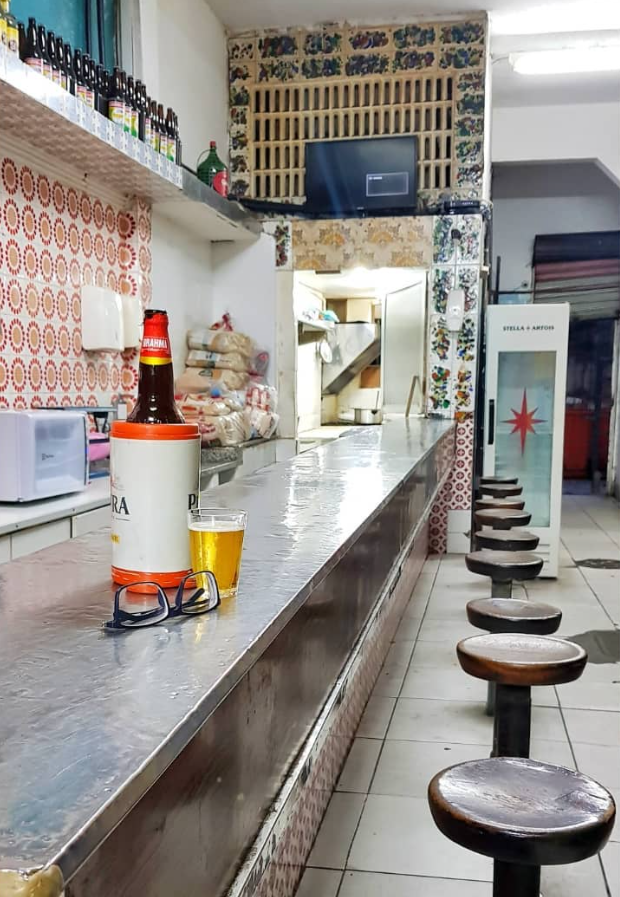 Foto de dentro do boteco, naquele balcão de metal, com os banquinhos um do lado do outro. Sobre o balcão, uma cerveja bem gelada