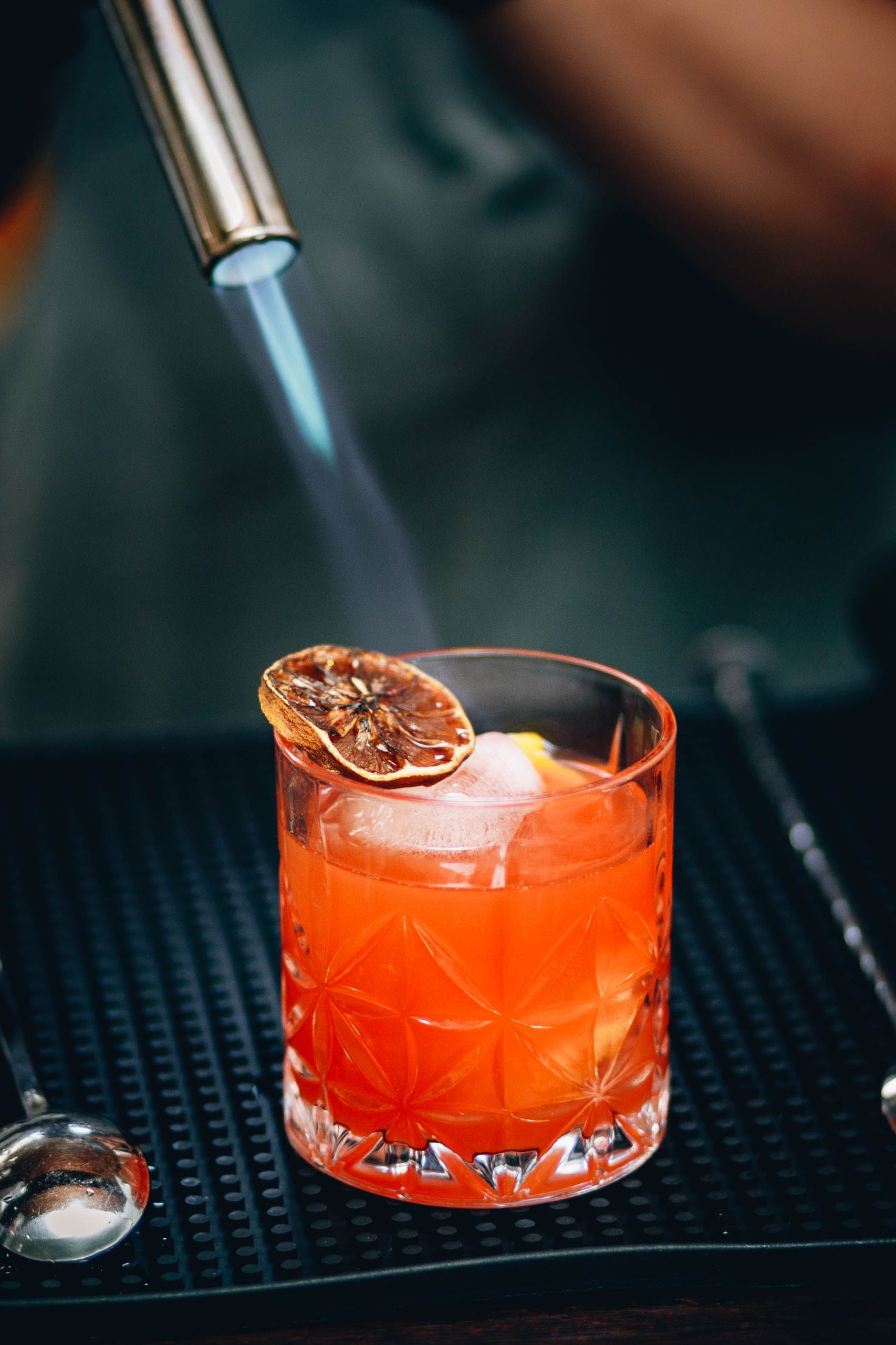 Foto de um drink laranja, com uma rodela de laranja sendo flambada sob o top do copo