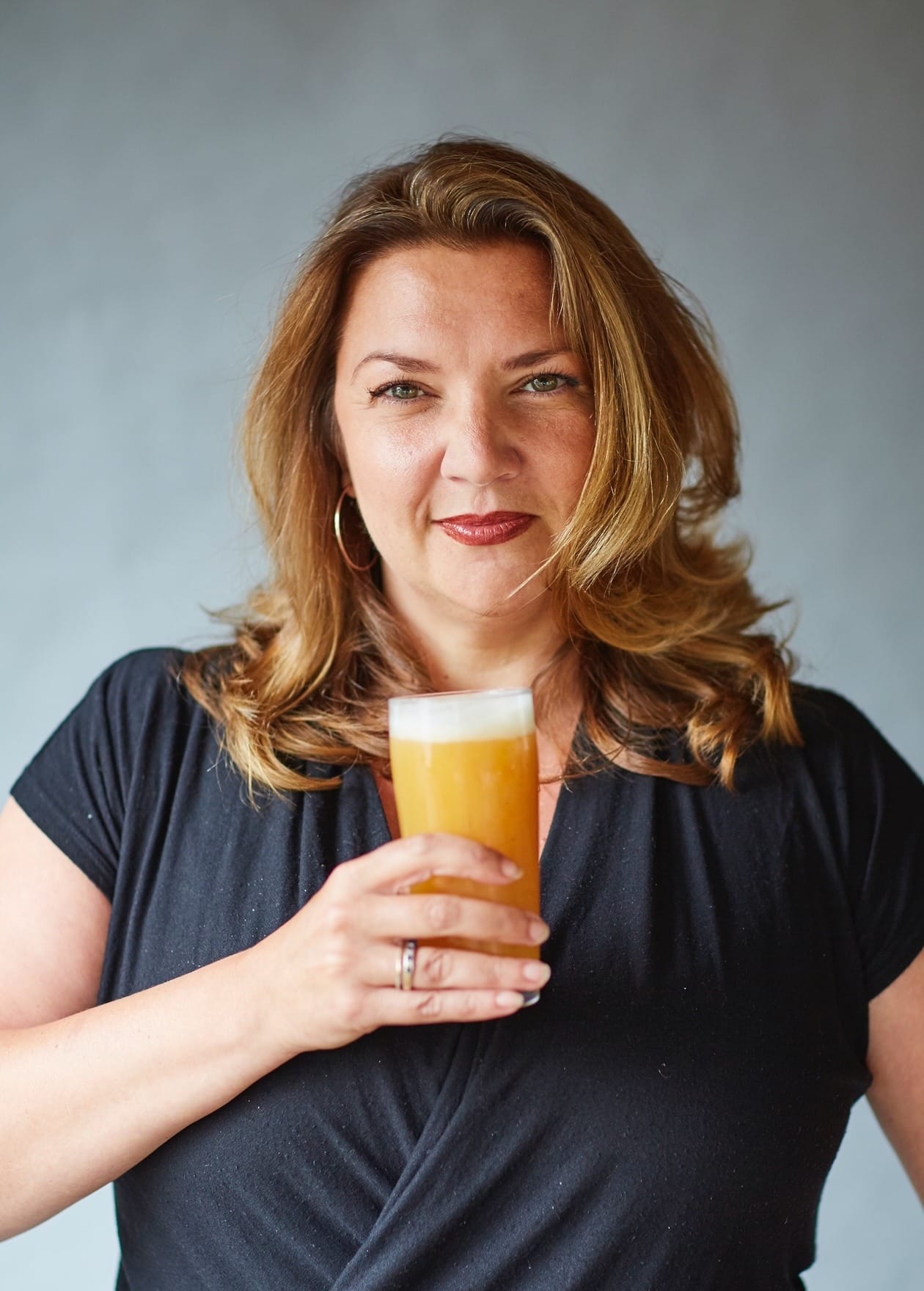Foto de Melissa Cole segurando cerveja. Mulher branca de camisa preta, segura copo de cerveja abaixo do queixo. Com cabelos loiros escuros, Melissa olha a câmera com um leve sorriso 